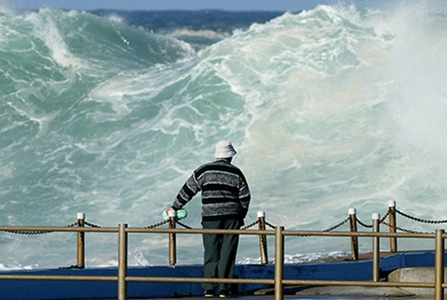 A bystander wearing warm clothing during winter stands on the edge of a saltwater swimming pool to watch surfers try to catch a large wave at Dee Why Point on Sydney’s north shore.